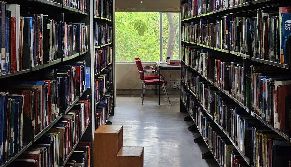 An open book glowing with golden light on a table in a grand multi-storey library of tall wooden bookshelves